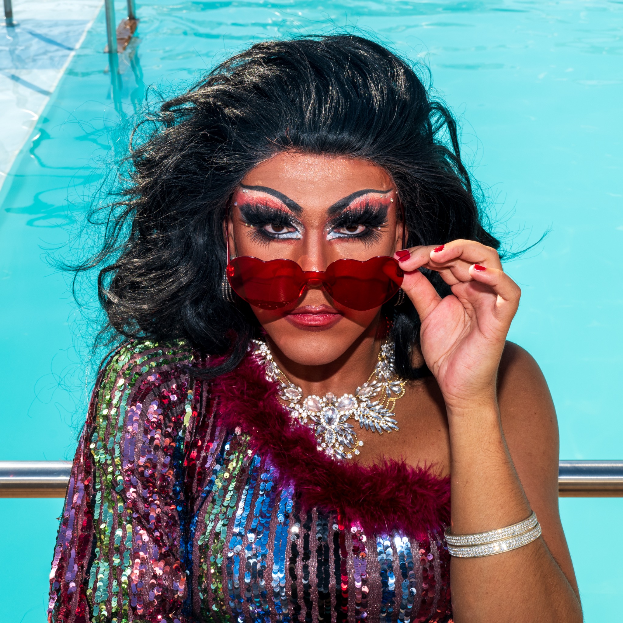Portrait in bright sequins and red sunglasses beside turquoise pool water.